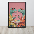 Kitchen wall art print featuring two stylized green dinosaurs wearing orange sunglasses and suits, sitting at a table with wine glasses against a pink background, displayed on a light wood floor against a white wall.