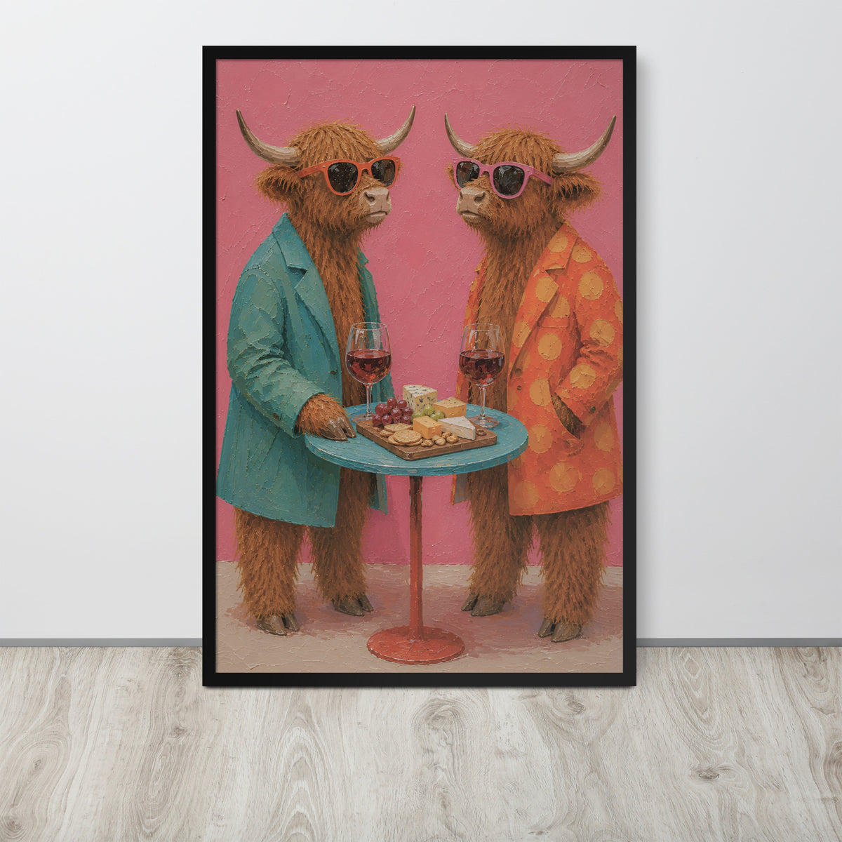 A framed funny farmhouse wall art print featuring two fluffy Highland cows in sunglasses, one in a teal blazer and one in an orange polka-dot jacket, standing behind a small table with wine glasses and cheese. The print is displayed on a light gray wall above a light wood floor.