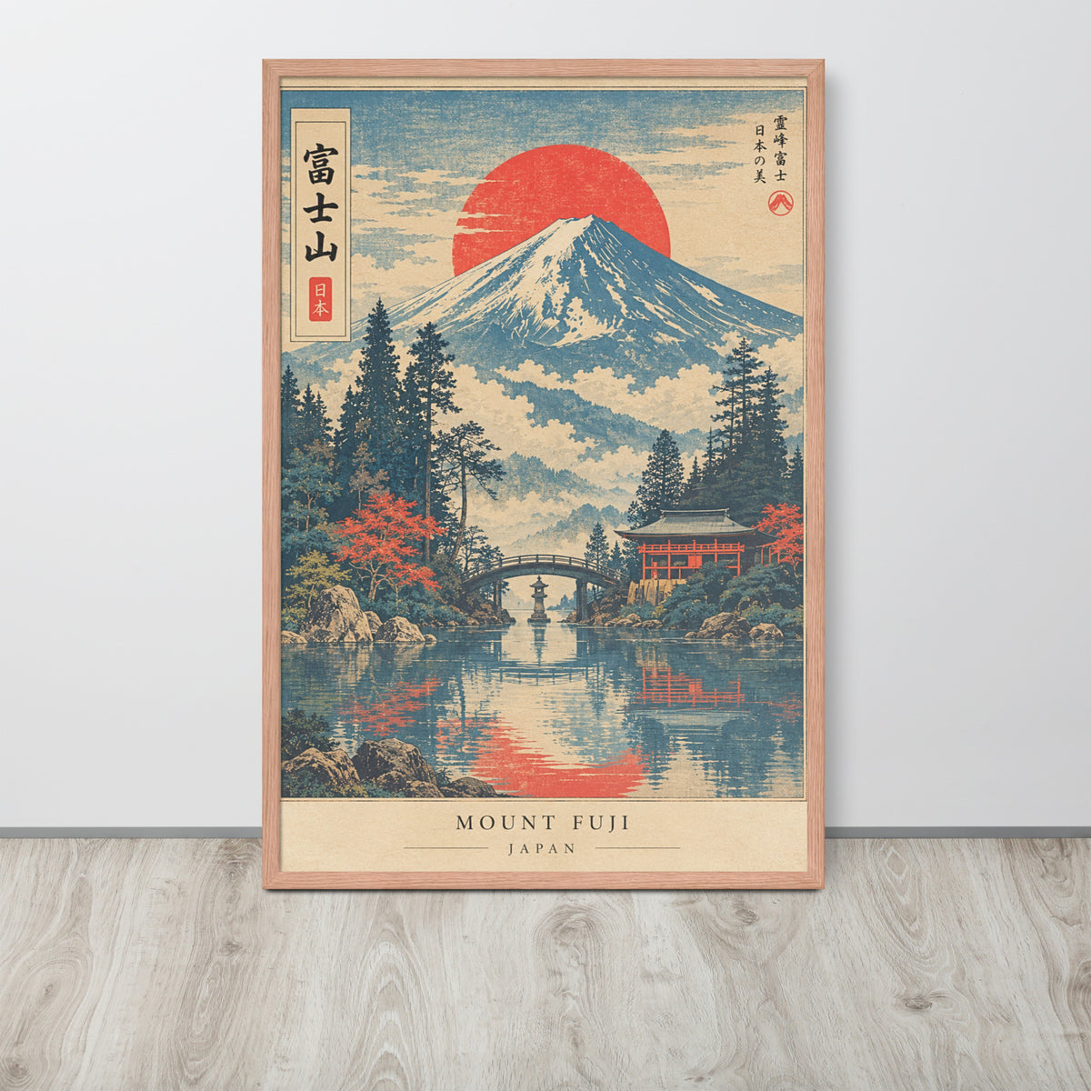 A framed Japanese-style art print of Mount Fuji with a large red sun, water reflection, trees, and a bridge, leaning against a light gray wall on a wood floor.