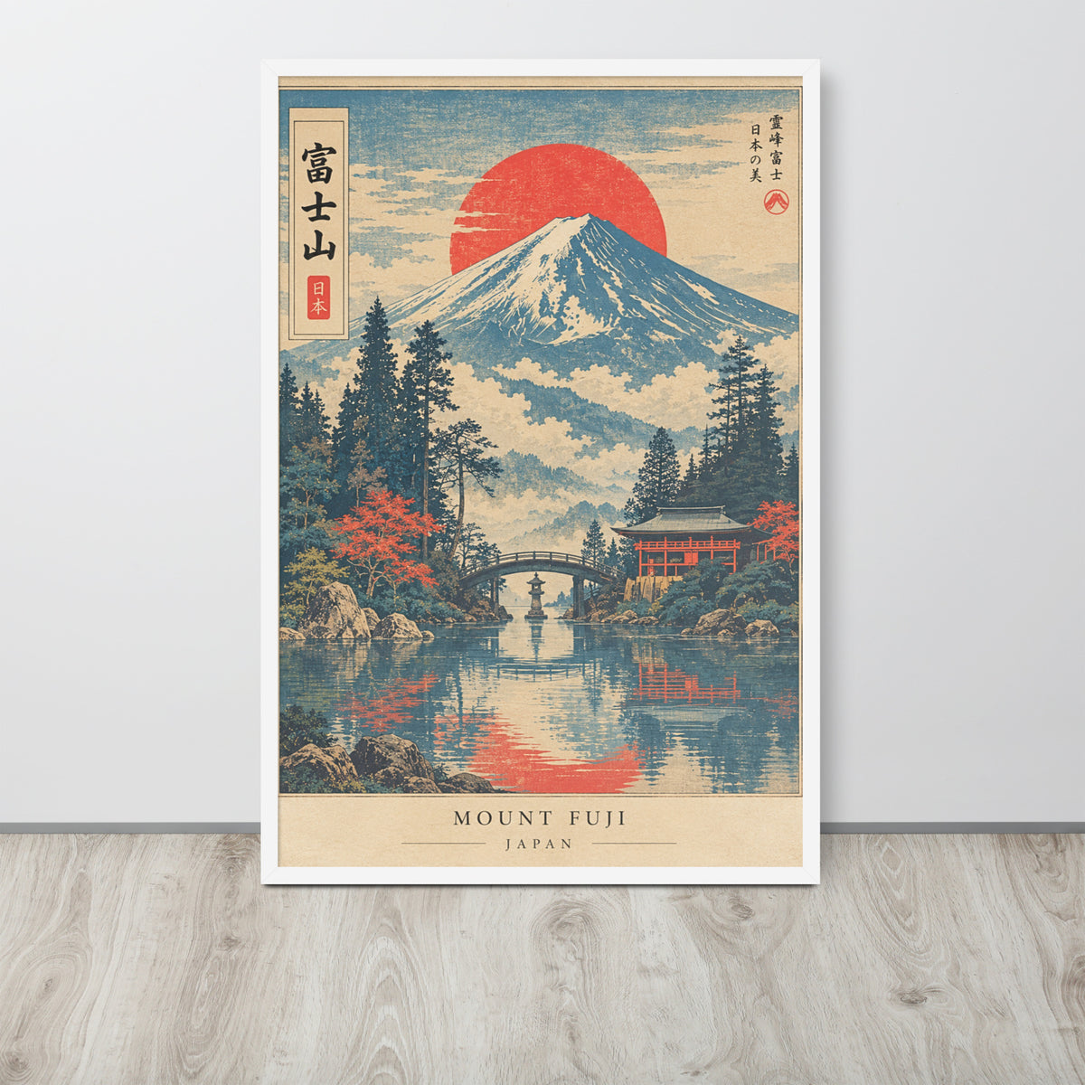 A framed art print of Mount Fuji in a traditional Japanese ukiyo-e style, leaning against a light gray wall on a light wood floor. The print features a large red sun, a snow-capped mountain, pine trees, and a red bridge over a reflective river.