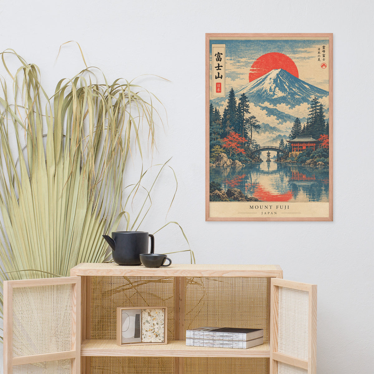 A framed Japanese-style art print of Mount Fuji with a red sun and reflective water, displayed on a light gray wall above a wooden shelf with a plant and black teapot in a styled interior setting.