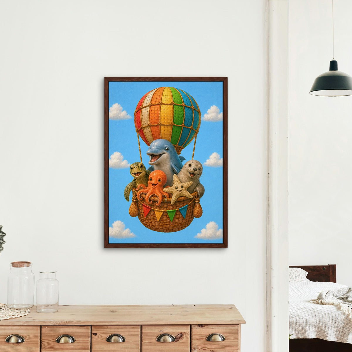 A framed nursery wall art print depicting a dolphin,  turtle, seal, starfish, and octopus in a hot air balloon basket with three kittens, displayed on a white wall in a modern bedroom setting.