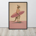 A framed Australian kangaroo wall art print featuring a kangaroo dressed as a surfer, holding a pink striped surfboard and wearing a striped shirt, standing on a skateboard against a beige and pink background. The print is displayed on a light gray wall above a light wood floor.