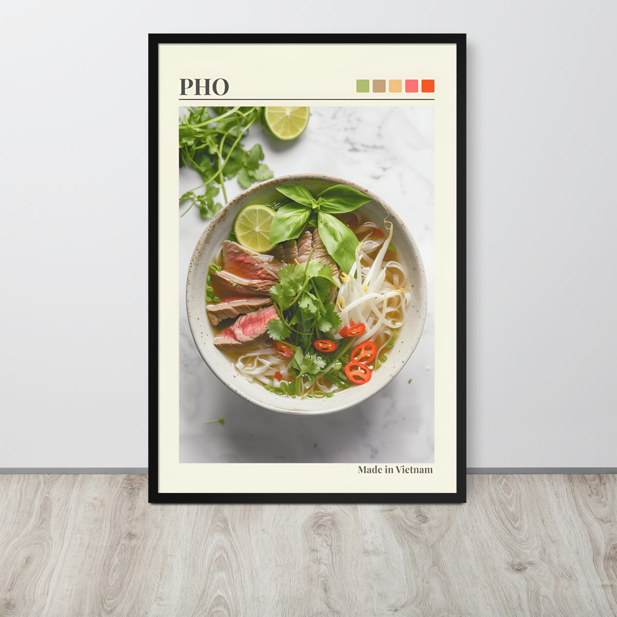 Pho Lover Decor art print featuring a bowl of pho noodle soup with basil, lime, and red chilli, displayed against a light wall on a wood floor. The print has a cream border, the word 'PHO' at the top, a small colour palette, and 'Made in Vietnam' at the bottom.